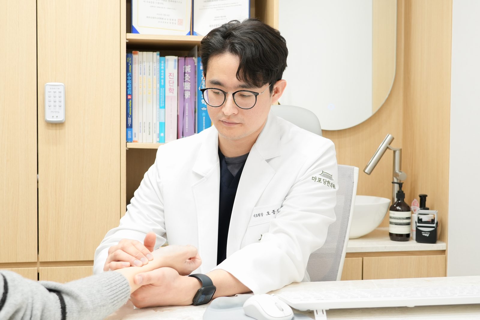 흰 가운을 입은 오종석 대표원장이 진료실 책상에서 환자의 손목 맥을 짚으며 소화불량 상태를 진찰하는 장면 - 마포당한의원
