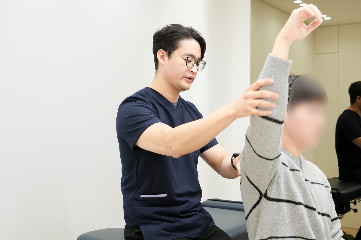 한의사가 환자의 어깨와 팔을 손으로 받쳐 주는 동안 환자가 직접 팔을 머리 위로 들어 올리는 멀리건 추나 시술 장면 - 마포당한의원